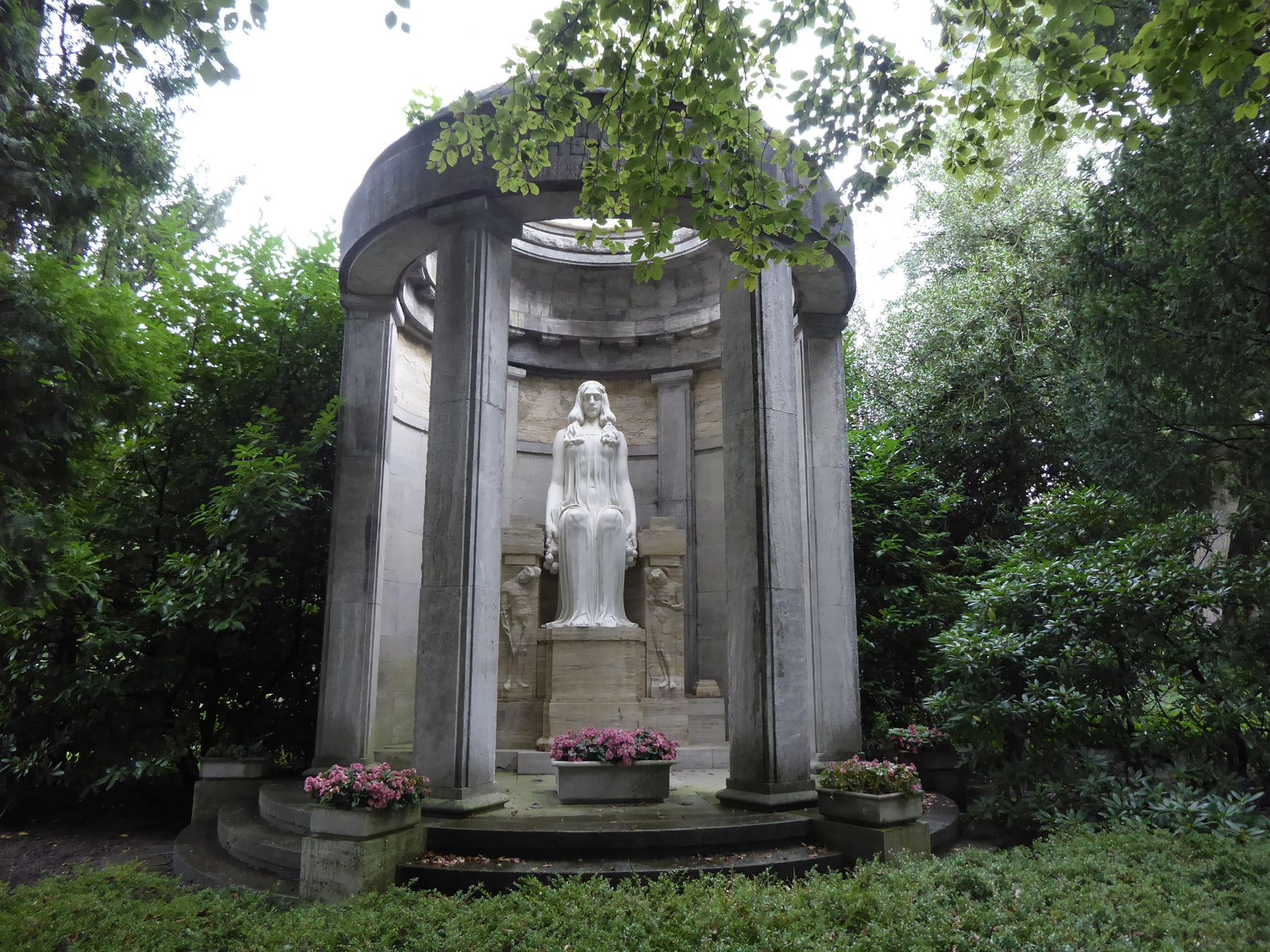 Runder Gedenkpavillon mit sitzender Frauenfigur und rosa Blumen zwischen Bäumen auf Nordfriedhof Düsseldorf